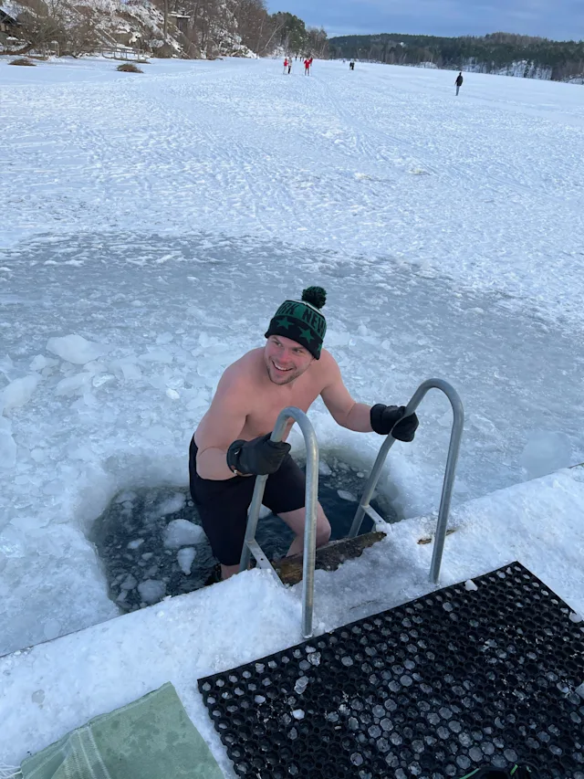 A person smiles while climbing out of an ice bath in a frozen lake. They wear a hat with NEW on it. Snowy landscape in the background.
