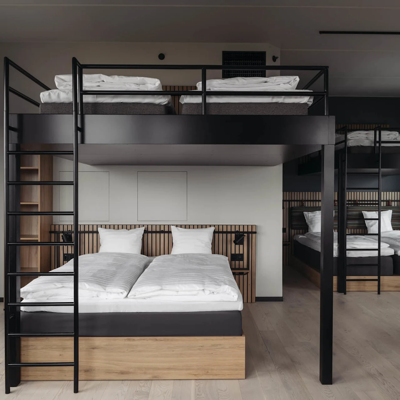 Two sets of bunk beds with wooden frames, neatly made with white linens, stand in a modern, minimalist room with light wood floors and minimal decor.