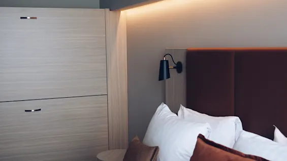 Et moderne hotellrom med en pent oppredd seng, myke puter, en stilig vegglampe og varm stemningsbelysning for et avslappende opphold.