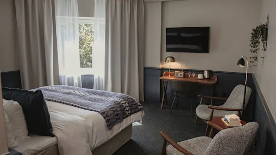 Ett mysigt hotellrum med en bekväm säng, en skrivbordshörna och två fåtöljer för avkoppling. Naturligt ljus fyller rummet.