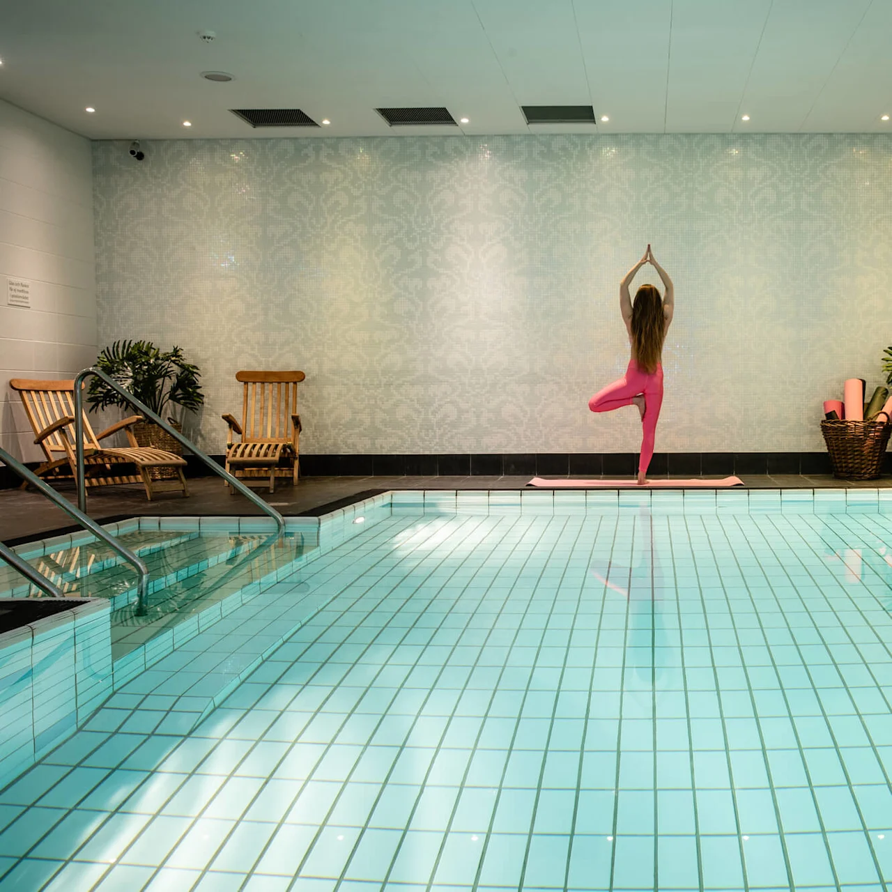 En person står i yogaposition vid en inomhuspool, omgiven av ljusblå väggar, växter, korgstolar och yogamattor. Skylten lyder: ”Varning, ursäkta eventuellt vatten på golvet.”