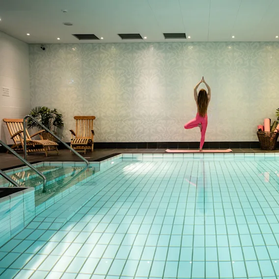En person står i yogaposition vid en inomhuspool, omgiven av ljusblå väggar, växter, korgstolar och yogamattor. Skylten lyder: ”Varning, ursäkta eventuellt vatten på golvet.”