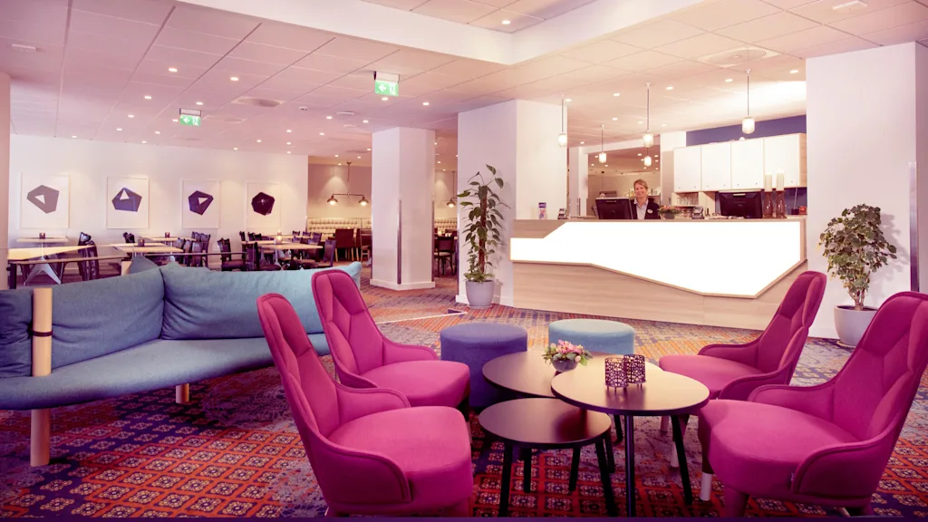 En lounge med soffor och fåtöljer i starka färger, en receptionsdisk och en person som står bakom den, i ett rymligt och välbelyst rum.
