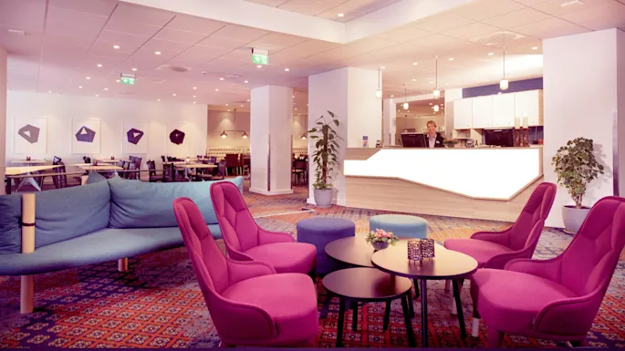 A lounge area with sofas and armchairs in vibrant colors, a reception desk, and a person standing behind it, set in a spacious, well-lit room.