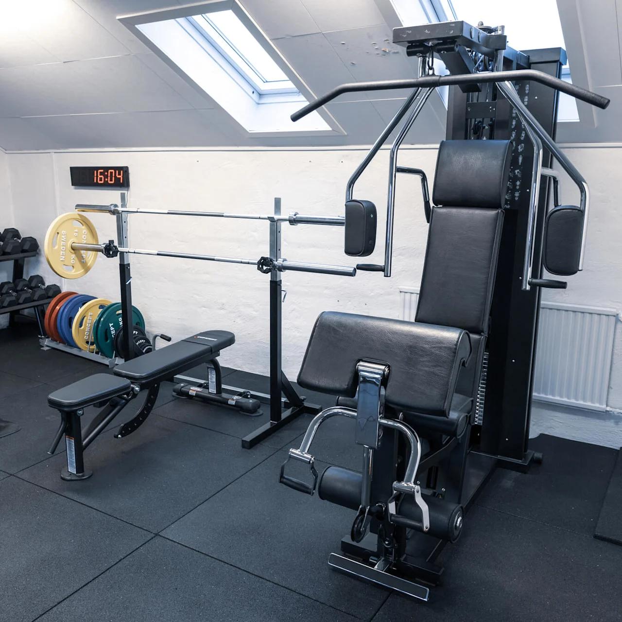 Et hjemmegym med vægtløftnings- og kabeltræningsudstyr, gummigulv, under et skråt tag med ovenlys.