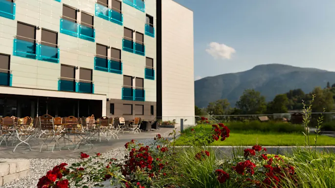 A modern building with blue-tinted balconies, surrounded by outdoor seating and red flowers, set against a backdrop of green hills and a clear sky.