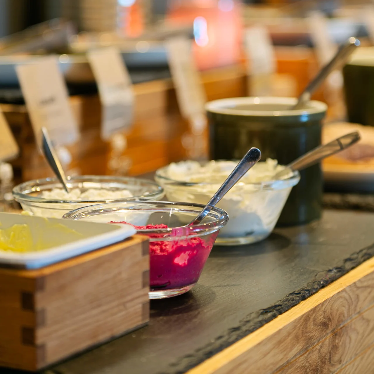 Skåle med farverige pålæg og skeer er placeret på et buffetbord, omgivet af forskellige krydderier og retter, i et spiseområde med sløret grønt i baggrunden.