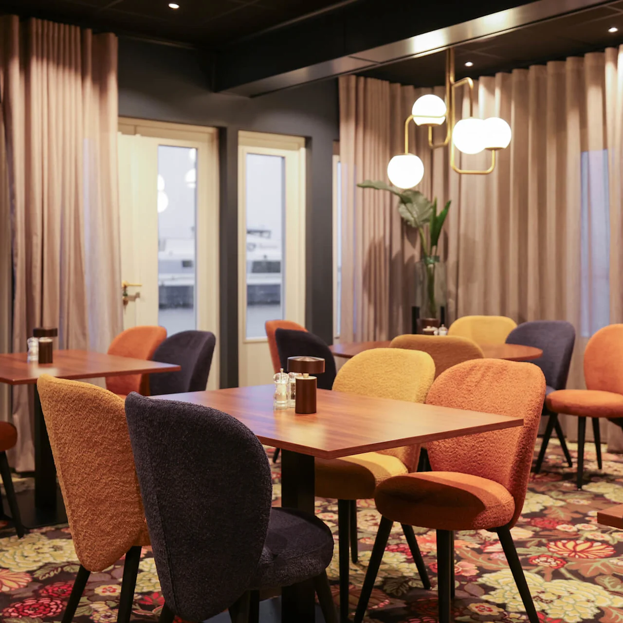 Colorful chairs surround wooden tables in a warmly lit room with large windows and floral-patterned carpet. Curtains cover some walls, with a hanging lamp creating a cozy atmosphere.