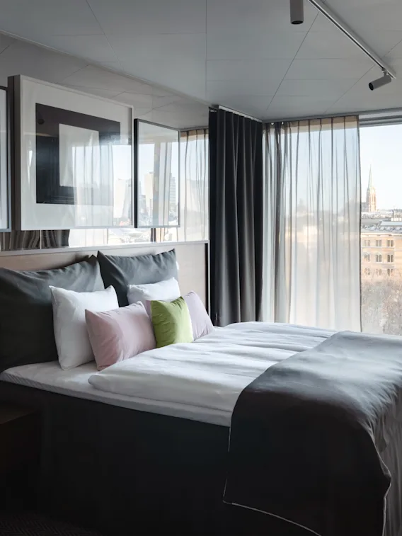 A modern hotel room with a comfortable bed, a stylish chair by a large window, and a city view.