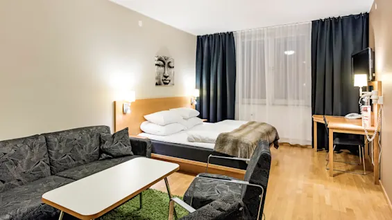 Et moderne hotellrom har en pent oppredd dobbeltseng med puter og et pledd, omgitt av gardiner, sofa, et lite bord, skrivebord og en veggmontert TV.