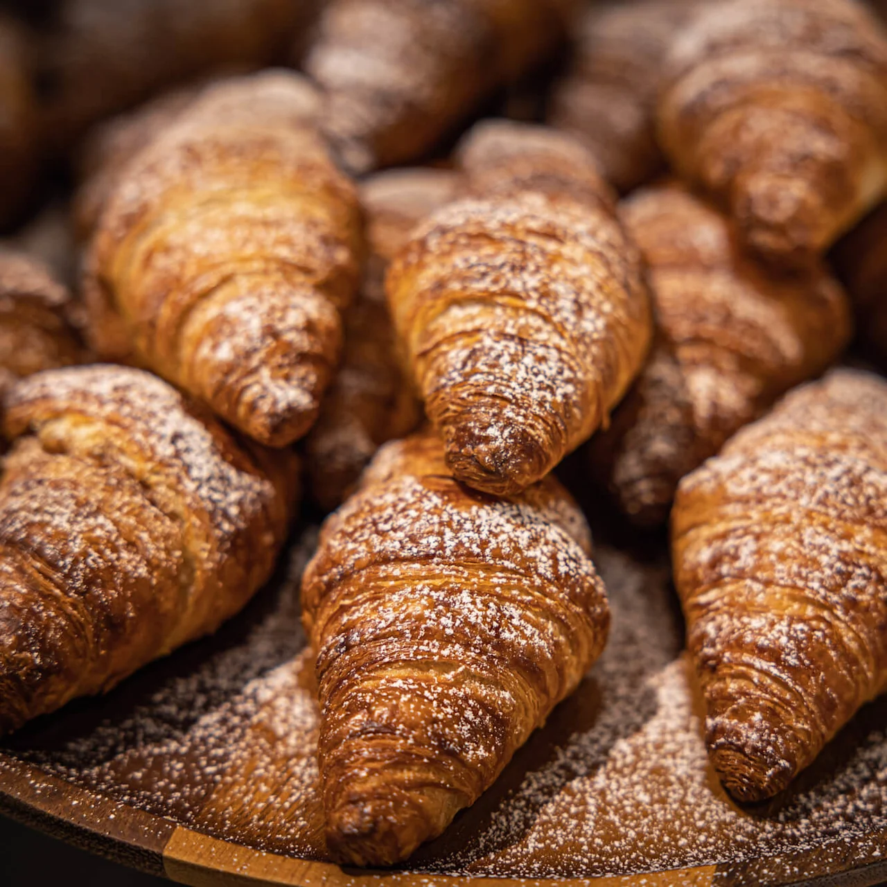 Croissanterne ligger tæt sammen på en træplade, drysset med flormelis, i en varm bageratmosfære.