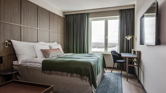 Moderne hotelværelse med en pænt opredt seng med grønt tæppe, skrivebord og stol nær et stort vindue med naturskøn udsigt. Træpaneler og en vægmonteret TV fuldender det hyggelige rum.