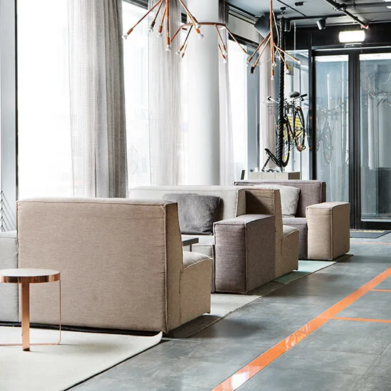 Gray and beige armchairs and sofas line a modern indoor lounge with large windows, sheer curtains, and contemporary lighting fixtures above. A bicycle is visible behind glass doors.