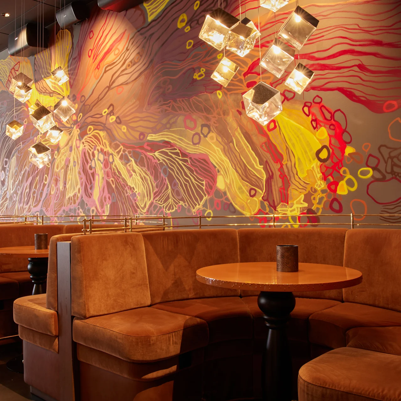 Bruna lädersoffor bildar en mysig matplats under glödande hänglampor, med en färgglad, abstrakt muralmålning som täcker väggen i en modern restaurangmiljö.