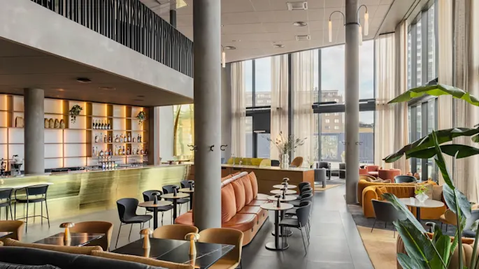 Et rummeligt, moderne loungeområde med stilfulde siddepladser og en elegant bar med hylder fyldt med drikkevarer. Store vinduer giver naturligt lys, mens planter og dekorationer skaber en indbydende atmosfære.