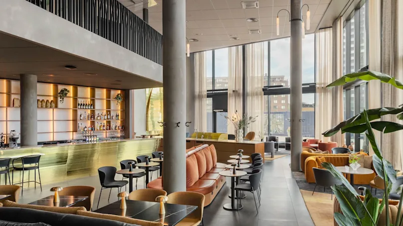 A spacious, modern lounge with stylish seating arrangements and a sleek bar with shelves of beverages. Large windows provide natural light, while plants and decor create a welcoming atmosphere.
