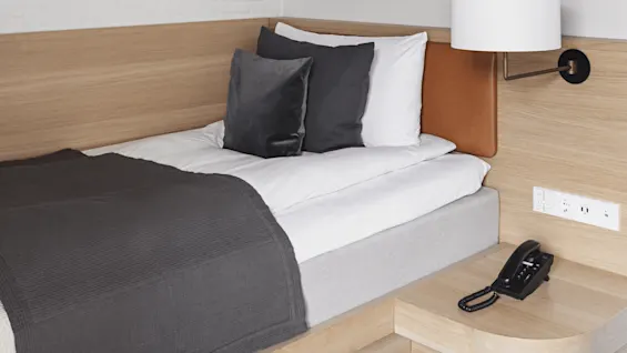 Bed with gray blanket and pillows, placed against white brick wall, under wooden beams. Bedside table holds a black telephone near power outlets and a wall-mounted lamp.