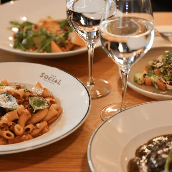 Pasta med sås och basilika på en tallrik märkt "SOCIAL", omgiven av andra rätter och två fyllda vinglas på ett träbord.