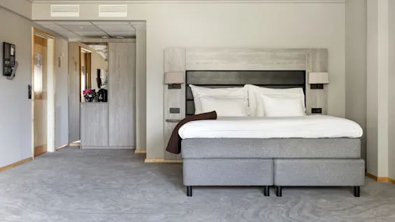A neatly made double bed with white linens and a brown throw rests against a wooden headboard in a carpeted hotel room. A hallway and minibar are visible.
