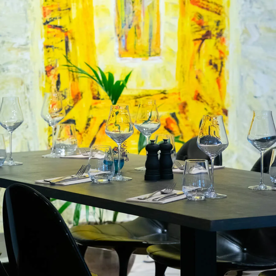 Several empty wine glasses are arranged on a dining table with utensils and salt shakers, set against a vibrant yellow abstract painting background. 