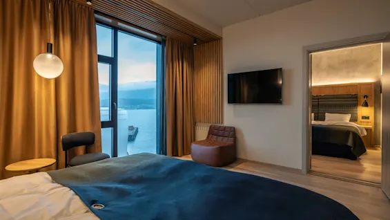 Et soveværelse med en seng dækket af et blåt tæppe; tilstødende siddeområde med stol, lille bord og moderne lampe; glasvindue med havudsigt; tilstødende rum synligt med TV.