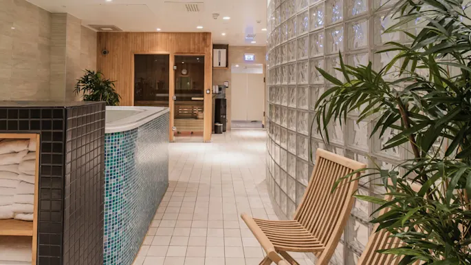 En tredør til en sauna ligger ved siden af et flisebelagt boblebad omgivet af planter, en træstol og glasblokvægge. Håndklæder er stablet i en hylde i nærheden.