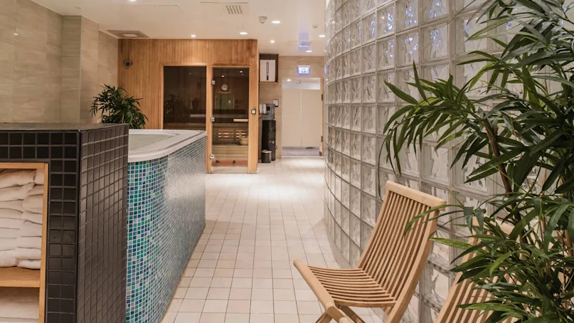 A wooden sauna door is adjacent to a tiled hot tub surrounded by plants, a wooden chair, and glass block walls. Towels are stacked nearby on a shelf.