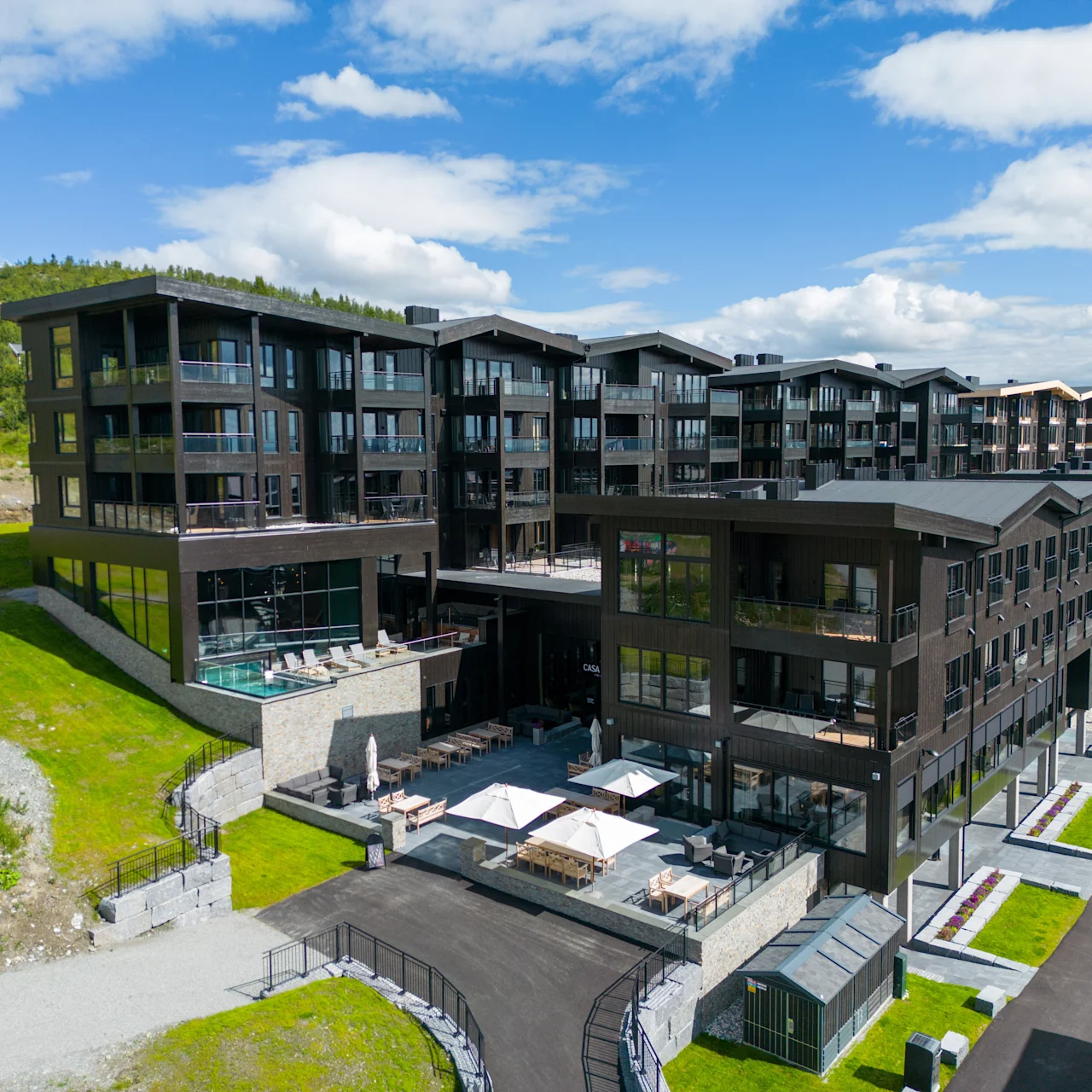 Et moderne, fler-etagers bygningskompleks med sorte facader står på en græsbeklædt skråning, med altaner og en rummelig terrasse. Omgivelser inkluderer klar himmel og nærliggende veje.