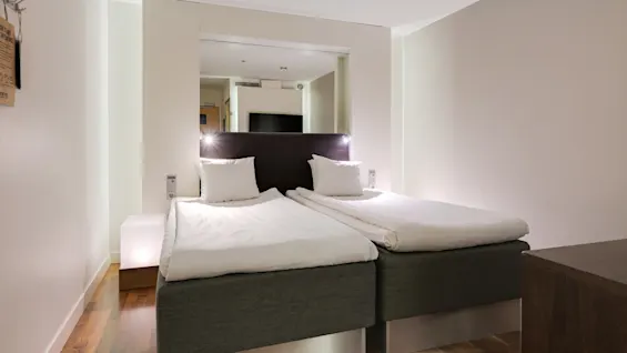 Två prydligt bäddade enkelsängar står bredvid varandra i ett modernt hotellrum. Ett trägolv och mjuk belysning skapar en varm, inbjudande atmosfär.