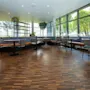 A modern, spacious cafeteria with wooden flooring and large windows, featuring tables and cushioned chairs arranged around the room. Indoor plants and natural light create a welcoming atmosphere.