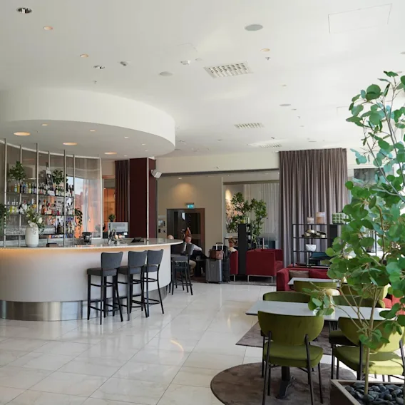 En modern lobbybar med en rundad disk, omgiven av stolar och frodiga växter. Människor sitter med bagage i bakgrunden, vilket tyder på en flygplats eller hotellmiljö.