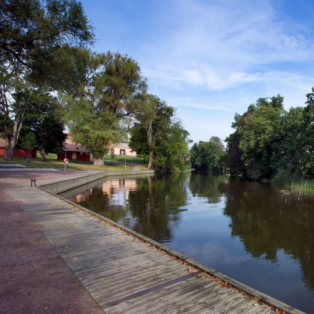A riverside with trees, a pedestrian, and a building reflecting on water in a serene urban setting.