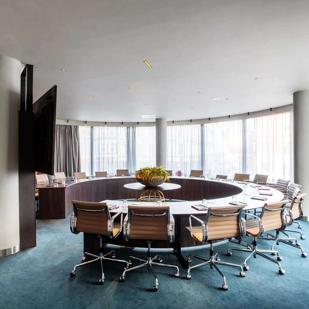 A round conference table is surrounded by office chairs, with a plant centerpiece, in a well-lit room with large windows and drapes, blue carpeting, and a wooden floor lamp.