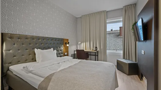 Ett mysigt hotellrum med en stor säng, ett funktionellt skrivbord vid fönstret och en väggmonterad TV. Rummet har en modern design med ljusa färger.