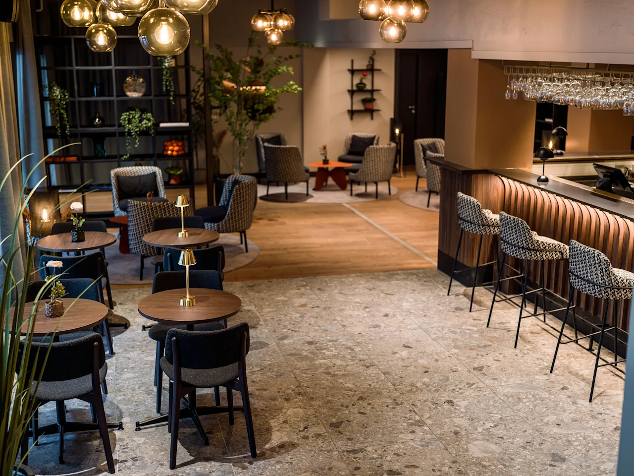 Mysig caféinteriör; runda bord med stolar, stämningsfull belysning ovanför, bar med pallar och växter skapar en inbjudande atmosfär. Hyllor och en liten lounge syns i bakgrunden.
