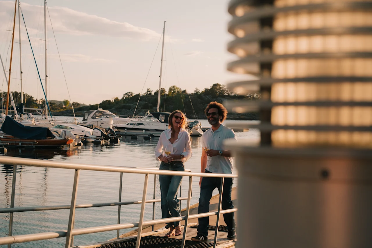 To personer nyder en gåtur på en kaj ved solnedgang, med både i baggrunden, holder drinks og smiler.