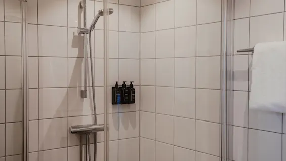 En modern hotalldusch med vita kakelplattor, ett duschhuvud och väggmonterade toalettartiklar. En fräsch vit handduk hänger i närheten.
