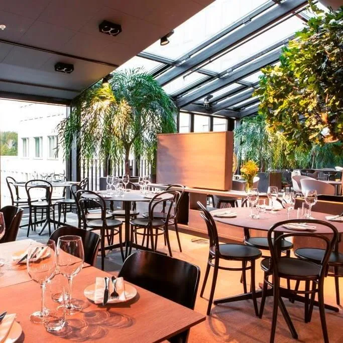 Restaurangbord är dukade med glas och bestick under ett glastak. Gröna växter omger sittgruppen, och stora fönster erbjuder en utsikt över ett skogsområde.