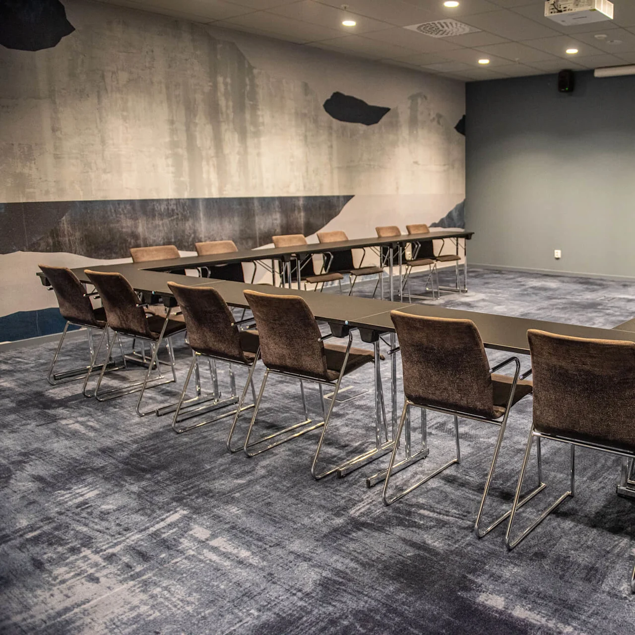 A large, modern conference room features a U-shaped arrangement of tables surrounded by chairs. The room has abstract wall art and a ceiling-mounted projector.