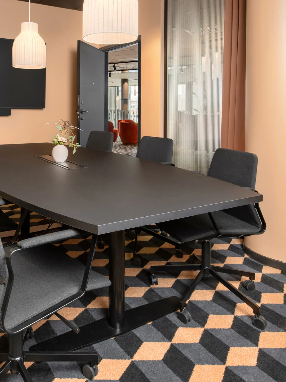 Ett stort svart konferensbord omgivet av sex stolar står i ett mötesrum med geometriskt mönstrad matta, väggmonterad skärm och hängande lampor ovanför.