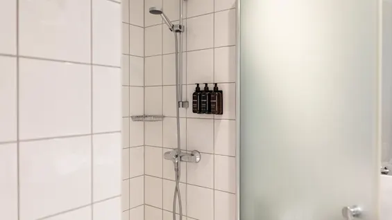 A clean, modern shower with white tiles, a chrome shower fixture, and three dark bottles of toiletries. A frosted glass door is visible.