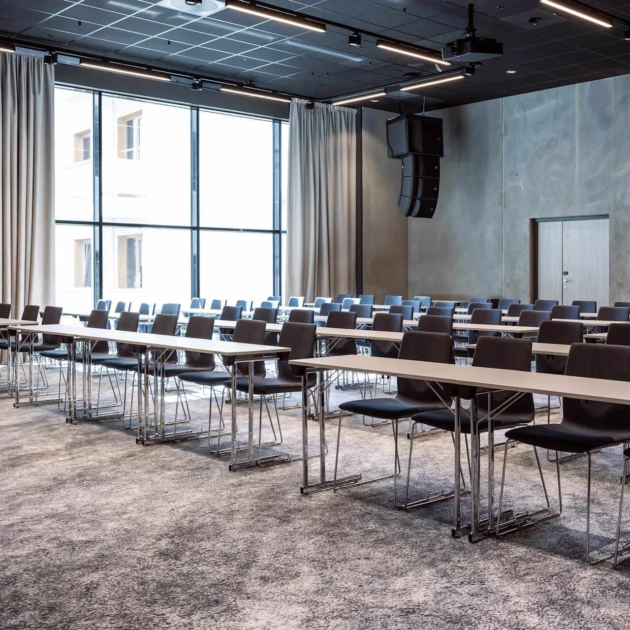 Rækker af tomme borde og stole fylder et moderne konferencelokale med store vinduer til venstre og en bølgemaleri på væggen til højre.