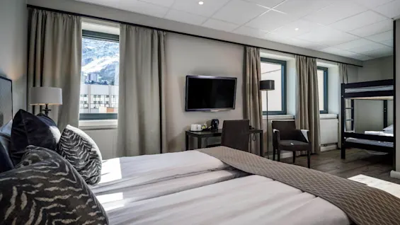 Et moderne hotelværelse har en stor seng med mønstrede puder, et vægmonteret tv, et lille bord, stole og en køjeseng. Vinduerne viser udsigt over snedækkede bjerge udenfor.