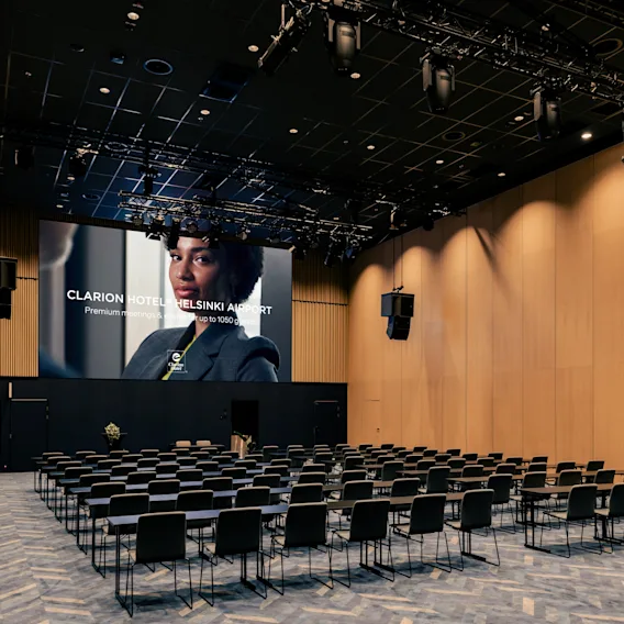 Konferenssihuone Clarion Hotel Helsinki Airportissa. Tuolit ja pöydät edessä näyttö: 'Hotel Helsinki Airport Premium meetings jopa 1050'.