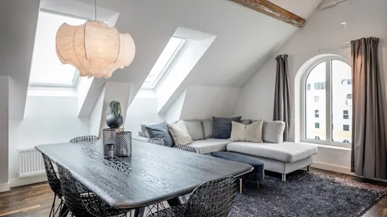 Ett mörkt träbord står under en stor, texturerad taklampa. I närheten står en grå sektionssoffa under lutande takfönster, med fönster från golv till tak och träbjälkar ovanför.