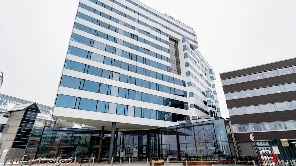 En moderne fleretasjesbygning med store glassvinduer ligger i et urbant område med flere bygninger i nærheten. Tekst på bygningen lyder: "CLARION HOTEL THE EDGE".