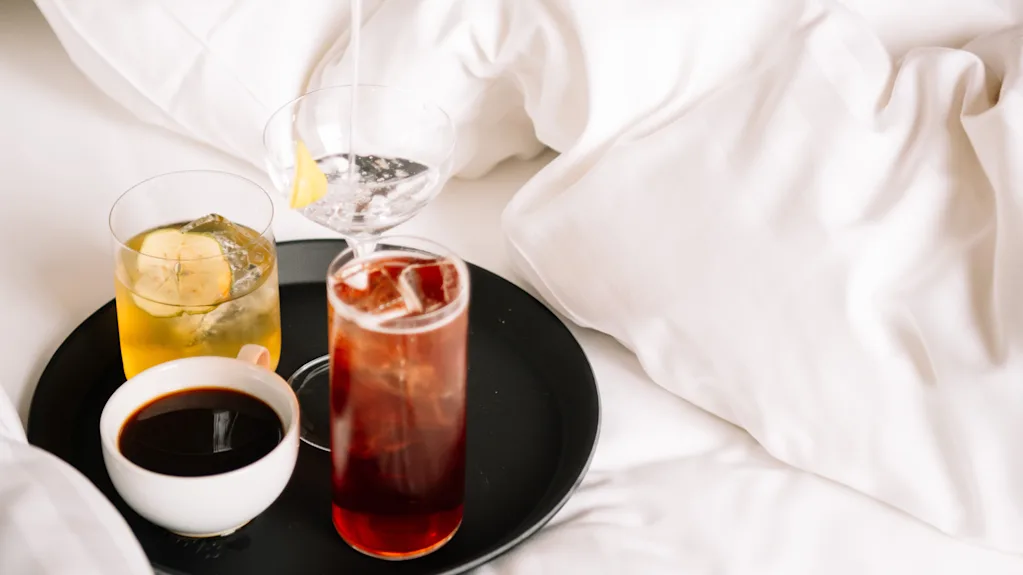 Käsi kaataa cocktailia ravistimesta lasiin valkoisella sängyllä olevalla tarjottimella.