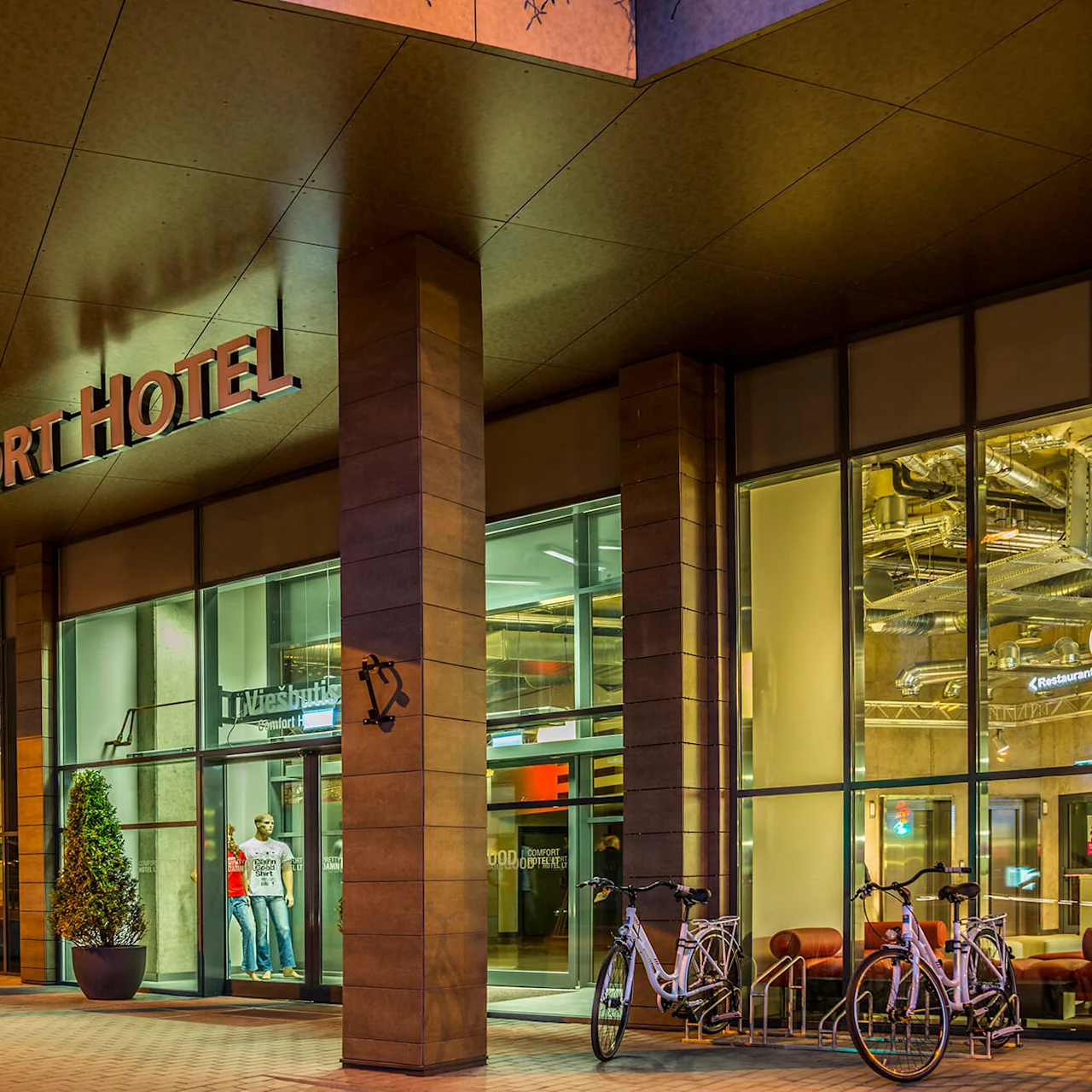 Entrén till ett Comfort Hotel, med cyklar parkerade utanför och en modern glasfasad. Skyltar lyder "Comfort Hotel" och "Restaurant." Exteriören är upplyst vid skymning.