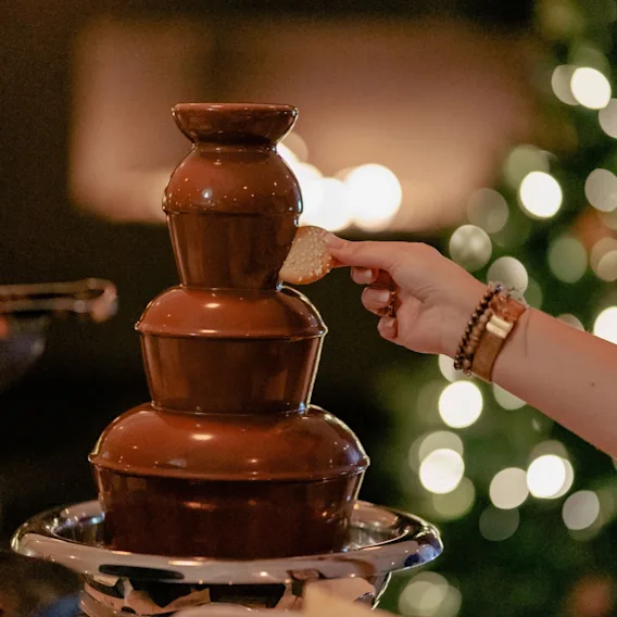 Nærbilde av en hånd som dypper en kjeks i en sjokoladefontene med varme julelys i bakgrunnen.