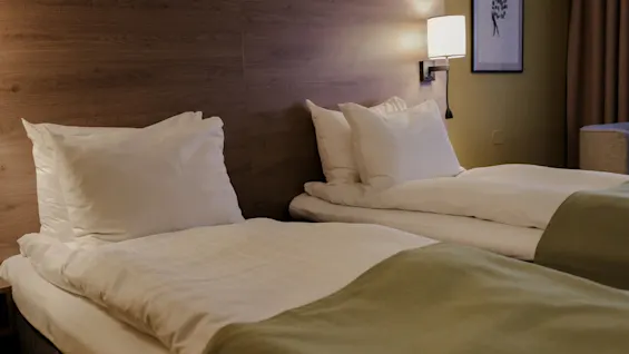 Two neatly made beds with white pillows and green blankets rest against a wooden wall, softly lit by a lamp. A framed picture hangs on the yellow wall.
